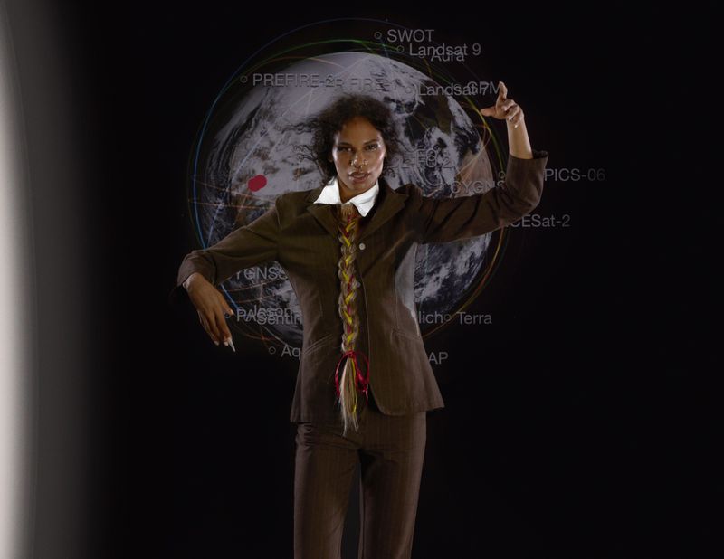Woman poses in a suit with braided hair, standing in front of a digital Earth projection with superimposed text in a darkened studio.