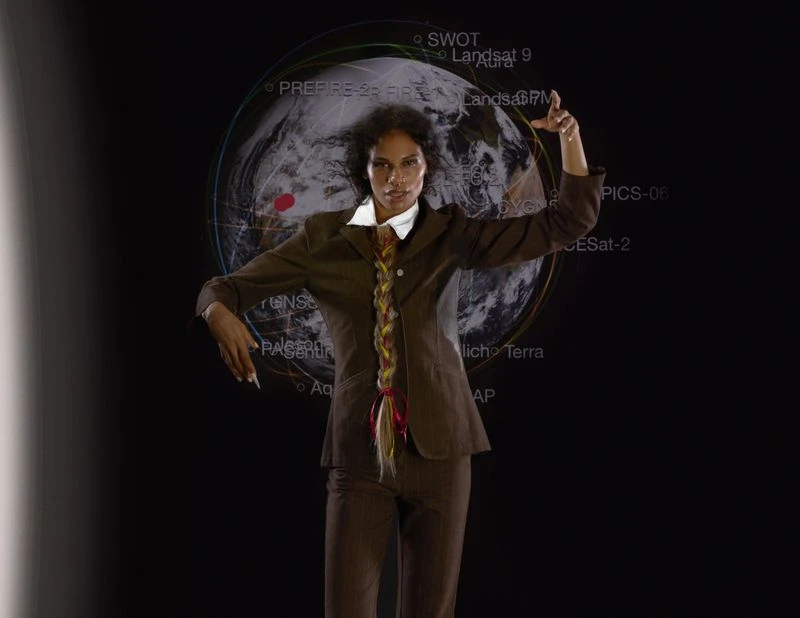 Woman poses in a suit with braided hair, standing in front of a digital Earth projection with superimposed text in a darkened studio.