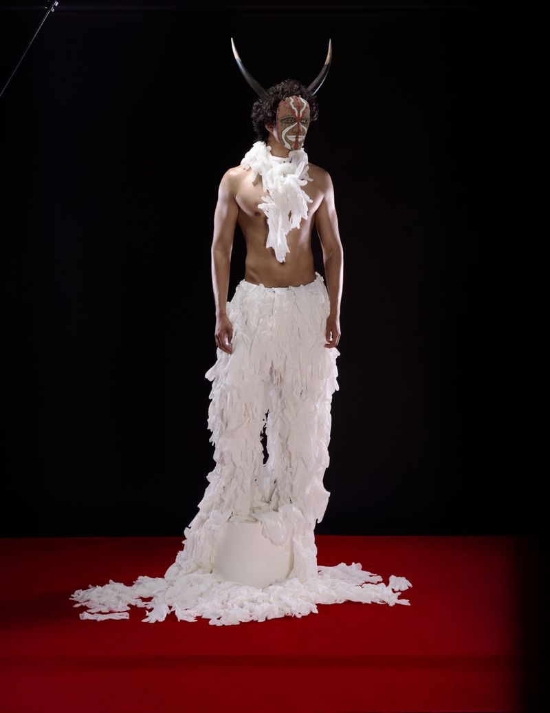 Adult male with white face paint, decorative shoulder piece, and large bull horns on head stands on a red surface in a studio.