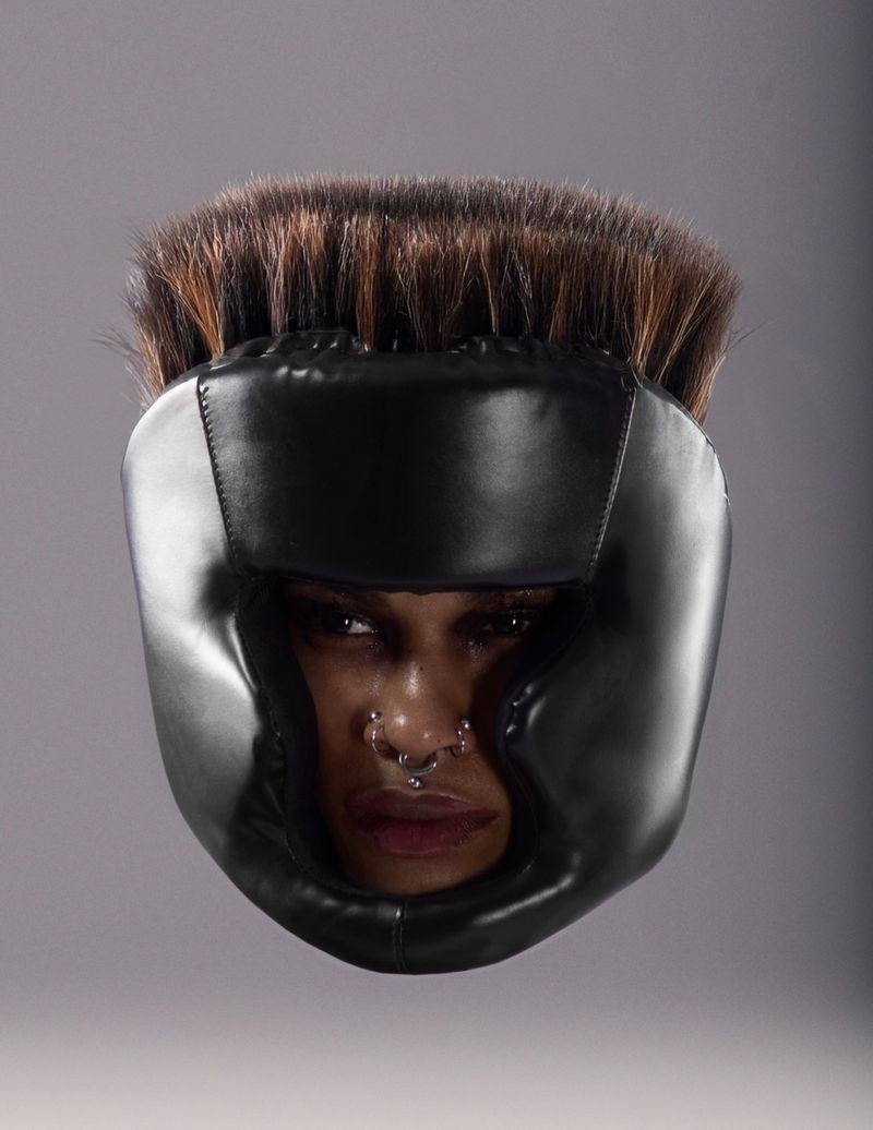 Woman wearing black boxing headgear with dark hair and a nose ring against a gray background in a studio shot, head and shoulders.