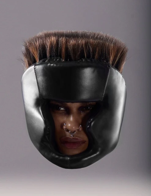 Woman wearing black boxing headgear with dark hair and a nose ring against a gray background in a studio shot, head and shoulders.