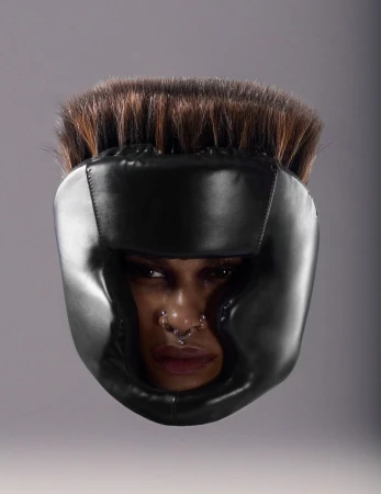 Woman wearing black boxing headgear with dark hair and a nose ring against a gray background in a studio shot, head and shoulders.