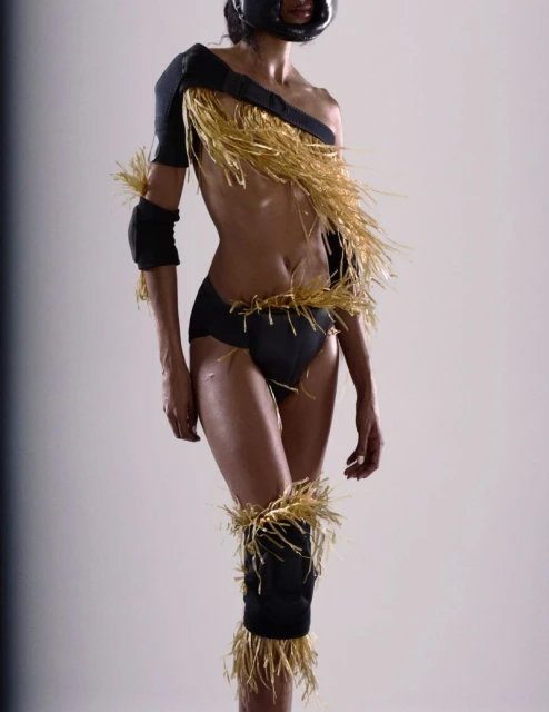 An athletic woman in black undergarments and football helmet poses in a studio, adorned with straw-like accents on her upper body and legs.