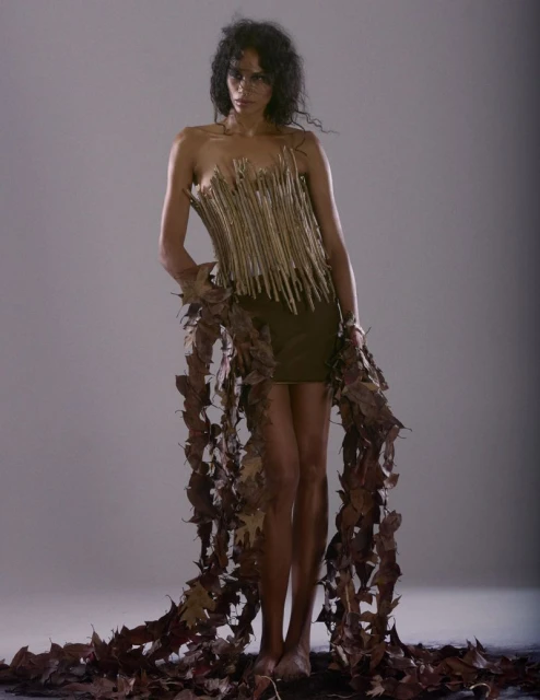 Adult female model poses in a strapless dress adorned with natural materials, including sticks and leaves, set against a neutral backdrop.