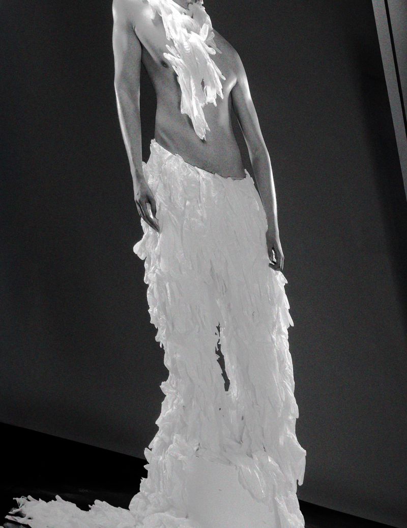 Fashion editorial featuring an adult model in a avant-garde white frilled costume against a dark gray background in an indoor setting.