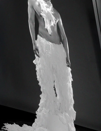Fashion editorial featuring an adult model in a avant-garde white frilled costume against a dark gray background in an indoor setting.