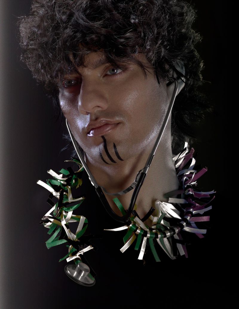 Headshot of a man with makeup and a stethoscope, suggesting a medical-themed artistic or conceptual portrait with dark background.