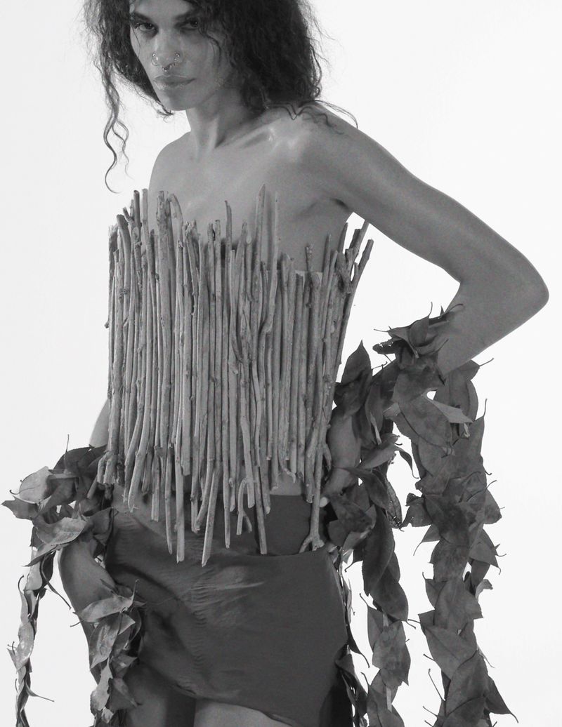 Black and white portrait of a person wearing a torso piece made of sticks and leaves in a minimalist studio setting.