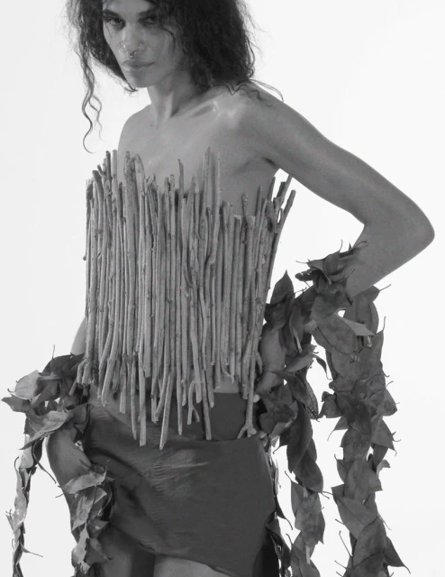 Black and white portrait of a person wearing a torso piece made of sticks and leaves in a minimalist studio setting.
