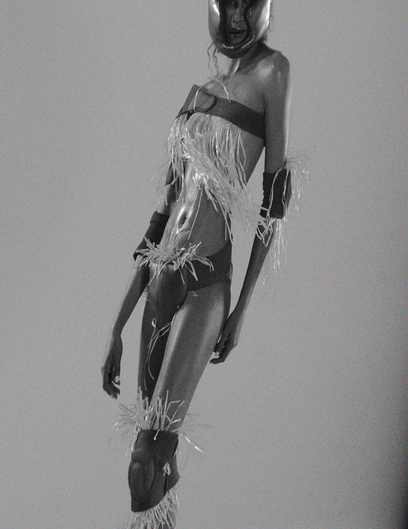Monochrome image of a model in a futuristic outfit with protective gear and feathers, posing in a studio with soft, even lighting.
