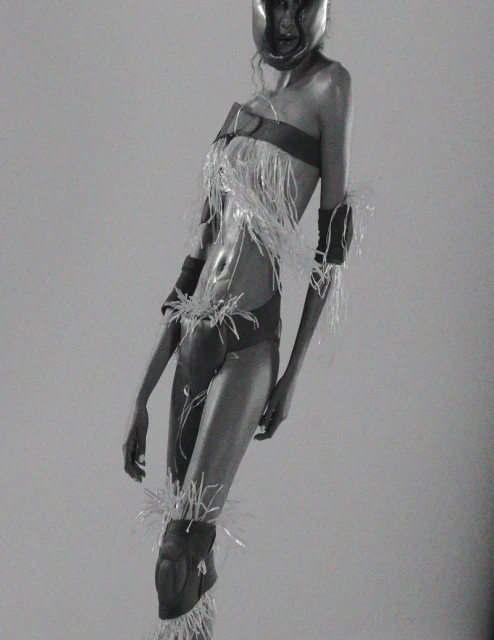 Monochrome image of a model in a futuristic outfit with protective gear and feathers, posing in a studio with soft, even lighting.