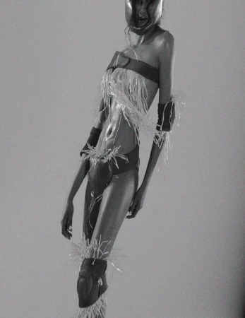 Monochrome image of a model in a futuristic outfit with protective gear and feathers, posing in a studio with soft, even lighting.