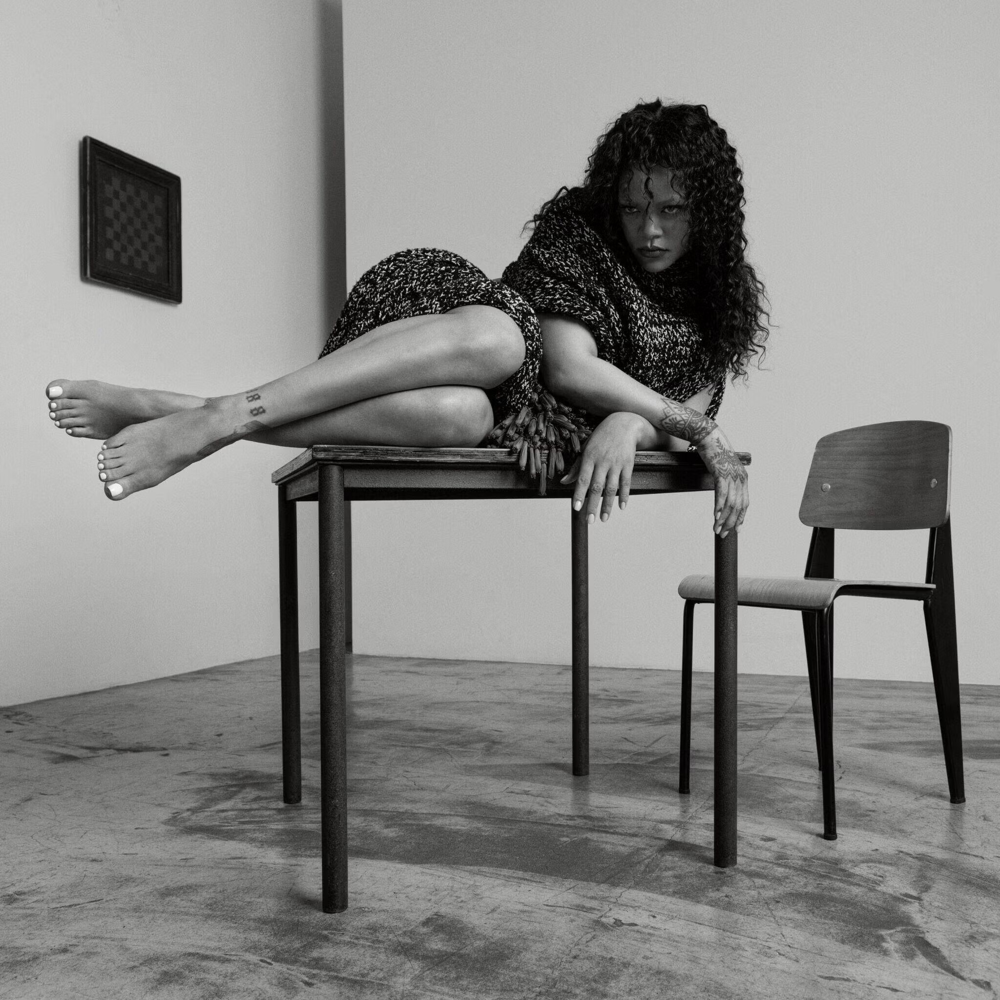Rihanna poses reclining on a dark table in a studio setting, wearing a textured dark garment with a matching wrap.