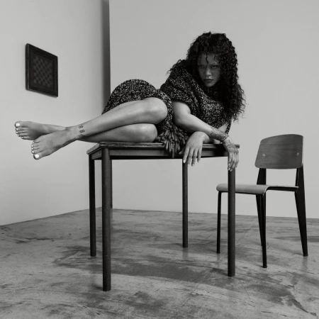 Rihanna poses reclining on a dark table in a studio setting, wearing a textured dark garment with a matching wrap.