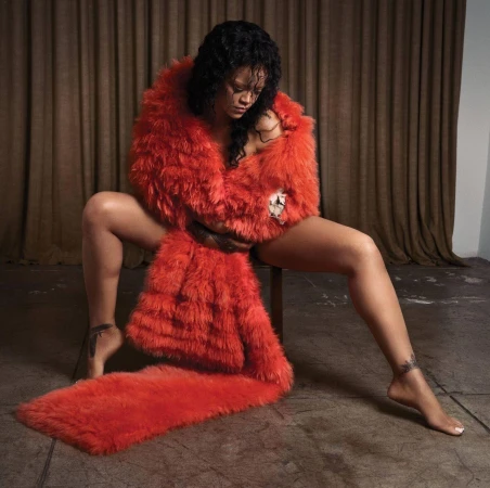 Rihanna sits on a chair wearing a vibrant red faux fur wrap that drapes to the floor, against a backdrop of brown curtains.