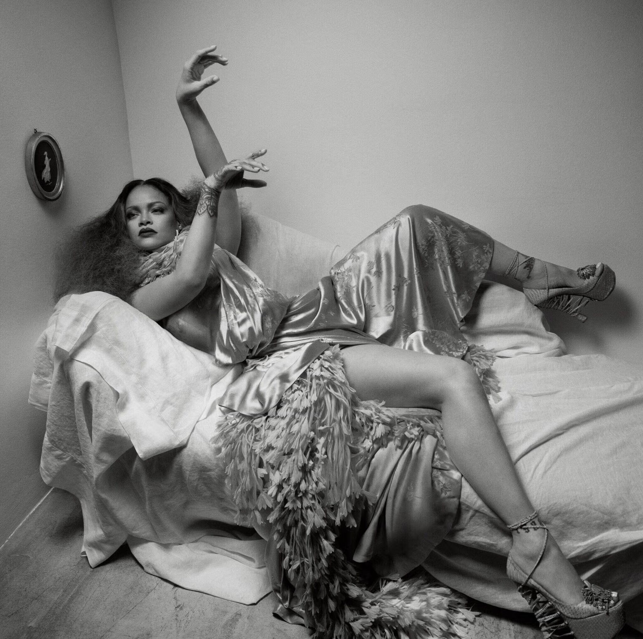 Rihanna reclines on a draped chaise lounge in a silk gown with a feather hem, arms raised, against a plain wall in this black and white portrait.