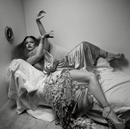 Rihanna reclines on a draped chaise lounge in a silk gown with a feather hem, arms raised, against a plain wall in this black and white portrait.