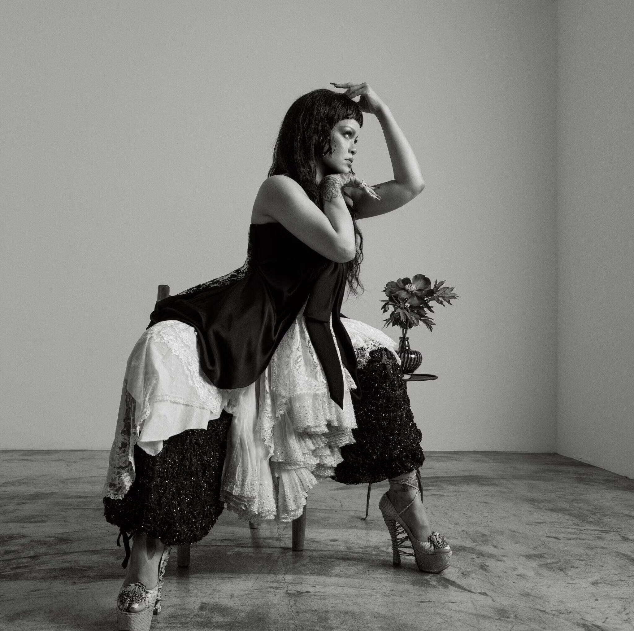 Rihanna seated in a chair, wearing a black satin top with lace-trimmed skirt and platform heels, posing against a white wall.