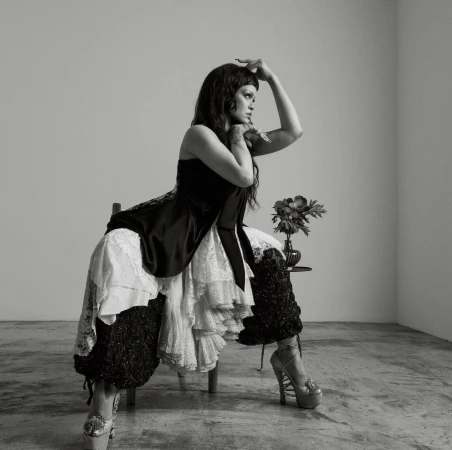 Rihanna seated in a chair, wearing a black satin top with lace-trimmed skirt and platform heels, posing against a white wall.