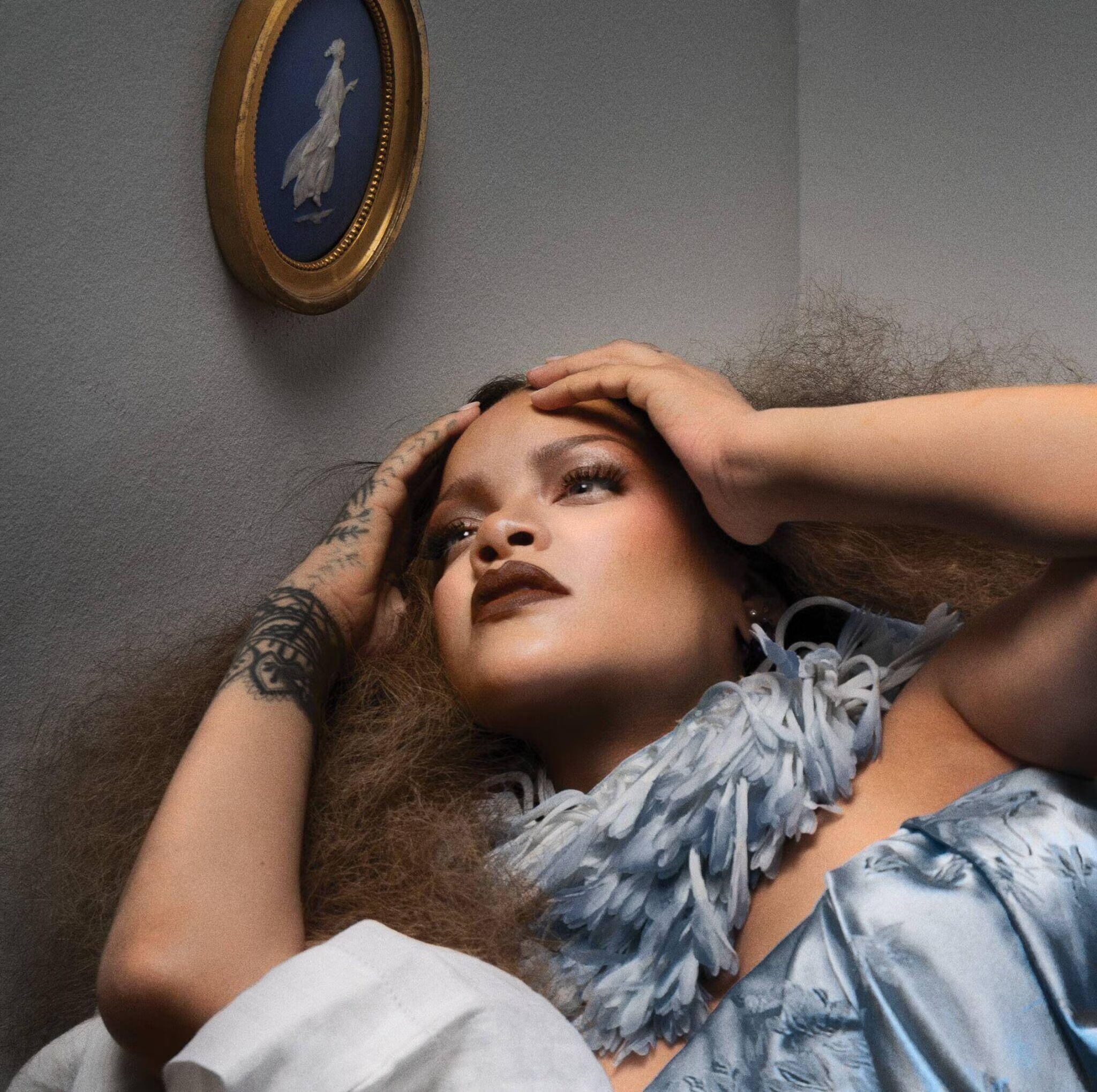 Rihanna wearing a blue textured collar with hands framing her face against a neutral wall with a blue and white cameo portrait.