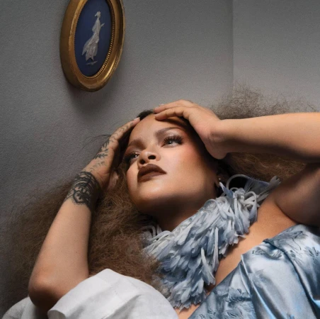 Rihanna wearing a blue textured collar with hands framing her face against a neutral wall with a blue and white cameo portrait.