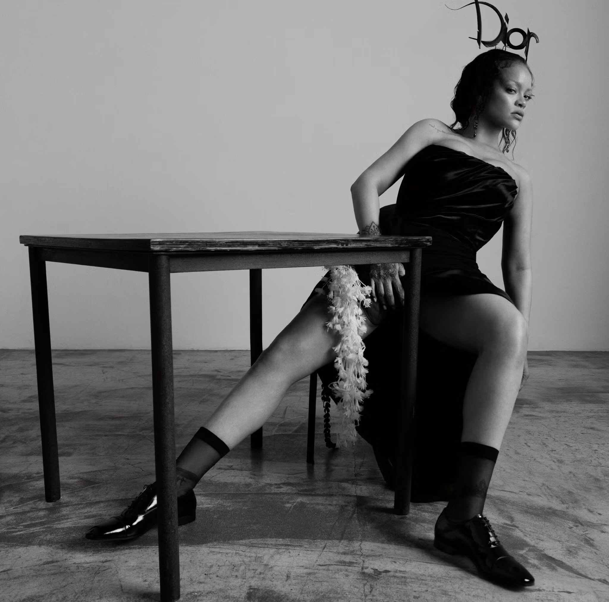 Rihanna wearing a black strapless dress and Dior headpiece seated at a wooden table in a studio, black and white portrait.