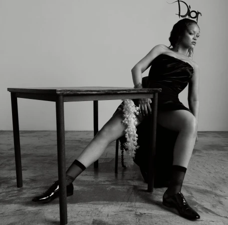 Rihanna wearing a black strapless dress and Dior headpiece seated at a wooden table in a studio, black and white portrait.