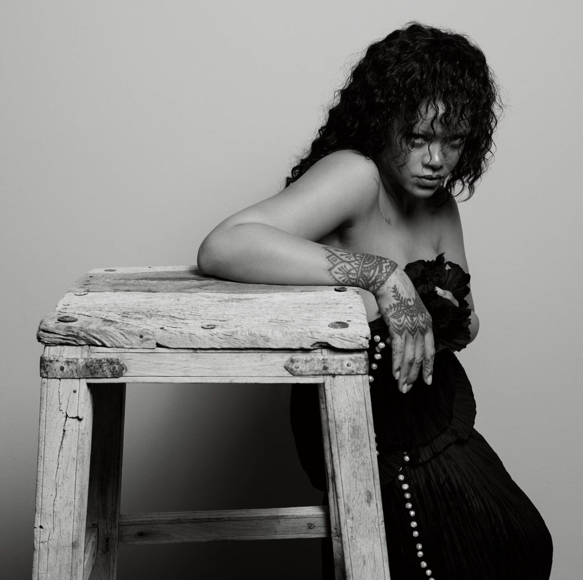 Rihanna posing against a rustic wooden stool, wearing a dark strapless dress, with visible henna-style hand and arm tattoos.