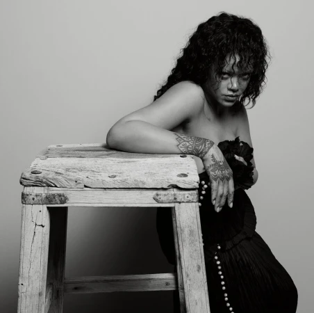 Rihanna posing against a rustic wooden stool, wearing a dark strapless dress, with visible henna-style hand and arm tattoos.