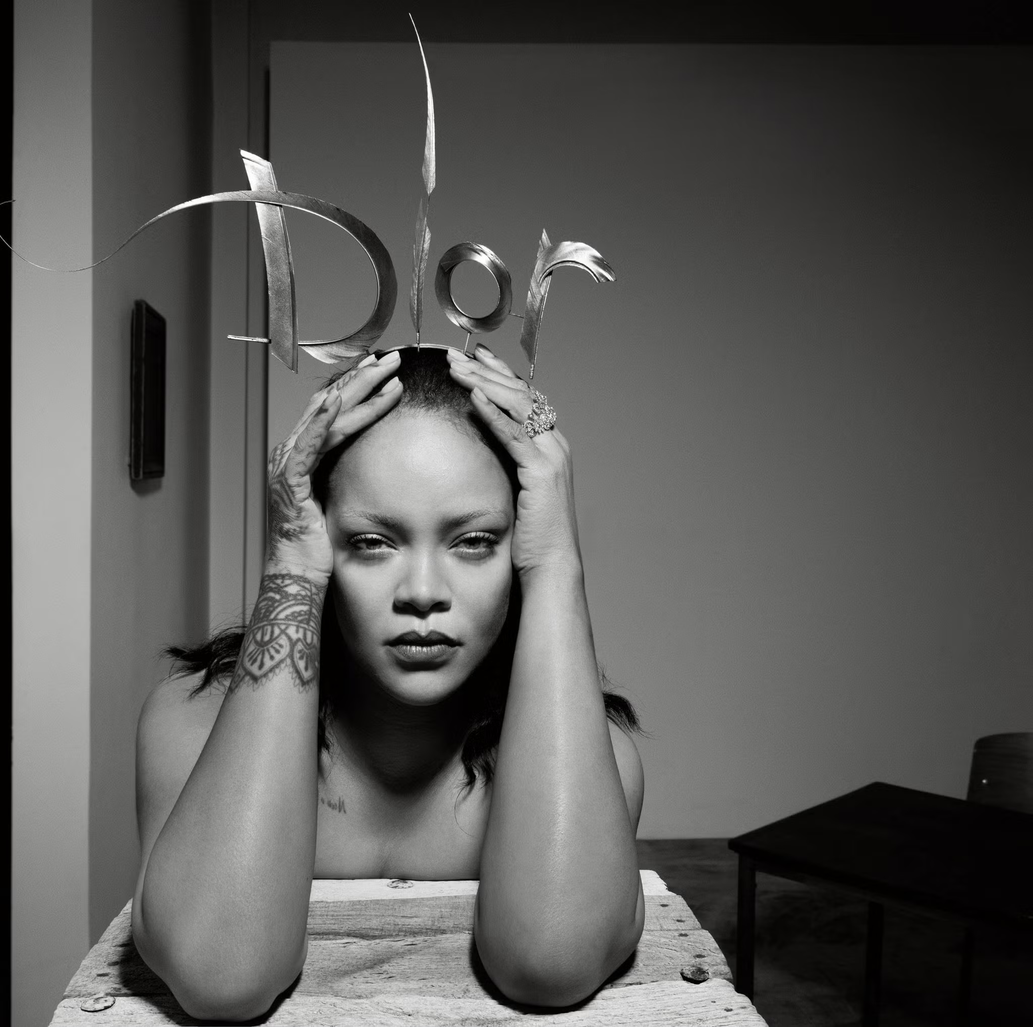 Rihanna poses with hands at her head wearing a silver Dior logo headpiece, against a minimalist grey background in black and white.