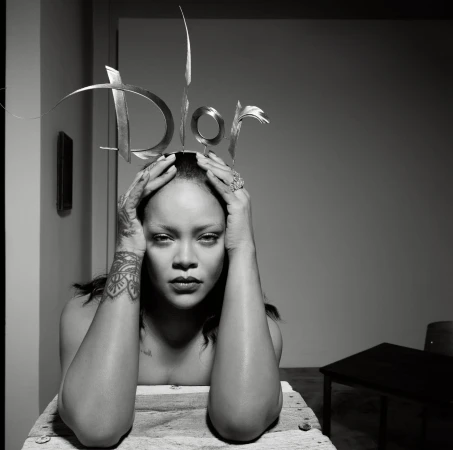 Rihanna poses with hands at her head wearing a silver Dior logo headpiece, against a minimalist grey background in black and white.