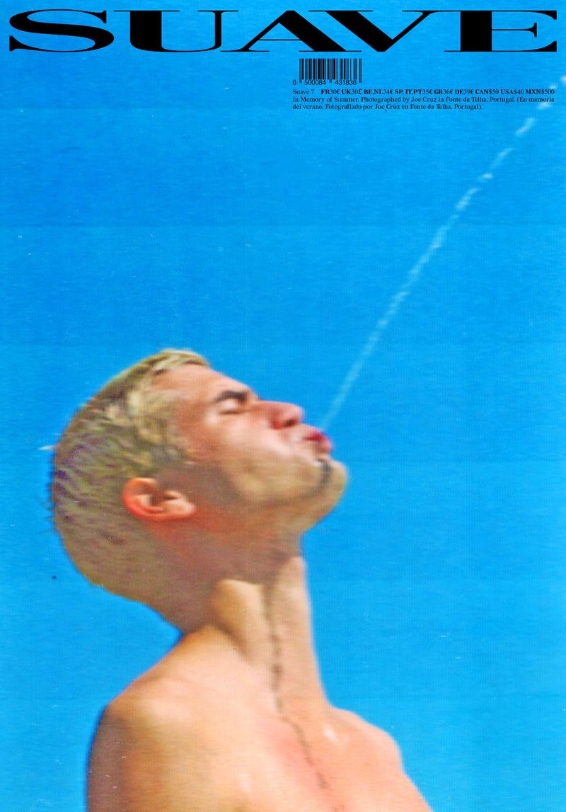 A close-up of a blonde-haired man with eyes closed against a bright blue sky, spitting a stream of water from his mouth into the air.