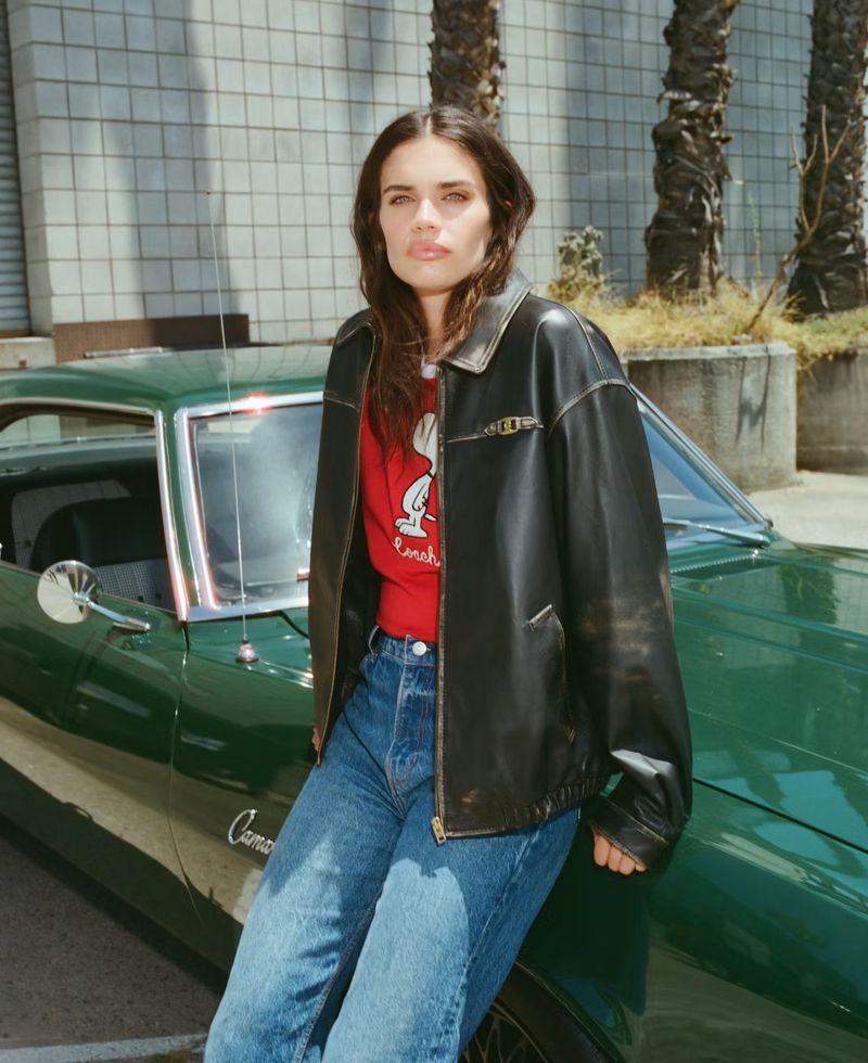 Brunette woman in black leather jacket poses by green vintage car outdoors in urban setting on sunny day.