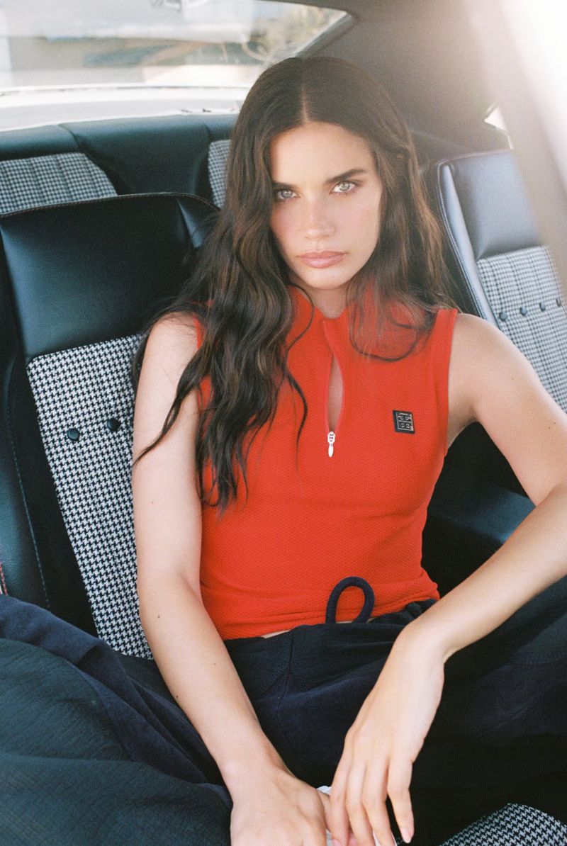 Adult woman sits in a classic car interior, wearing a red tank top and dark blue sweatpants, styled with wavy brunette hair.
