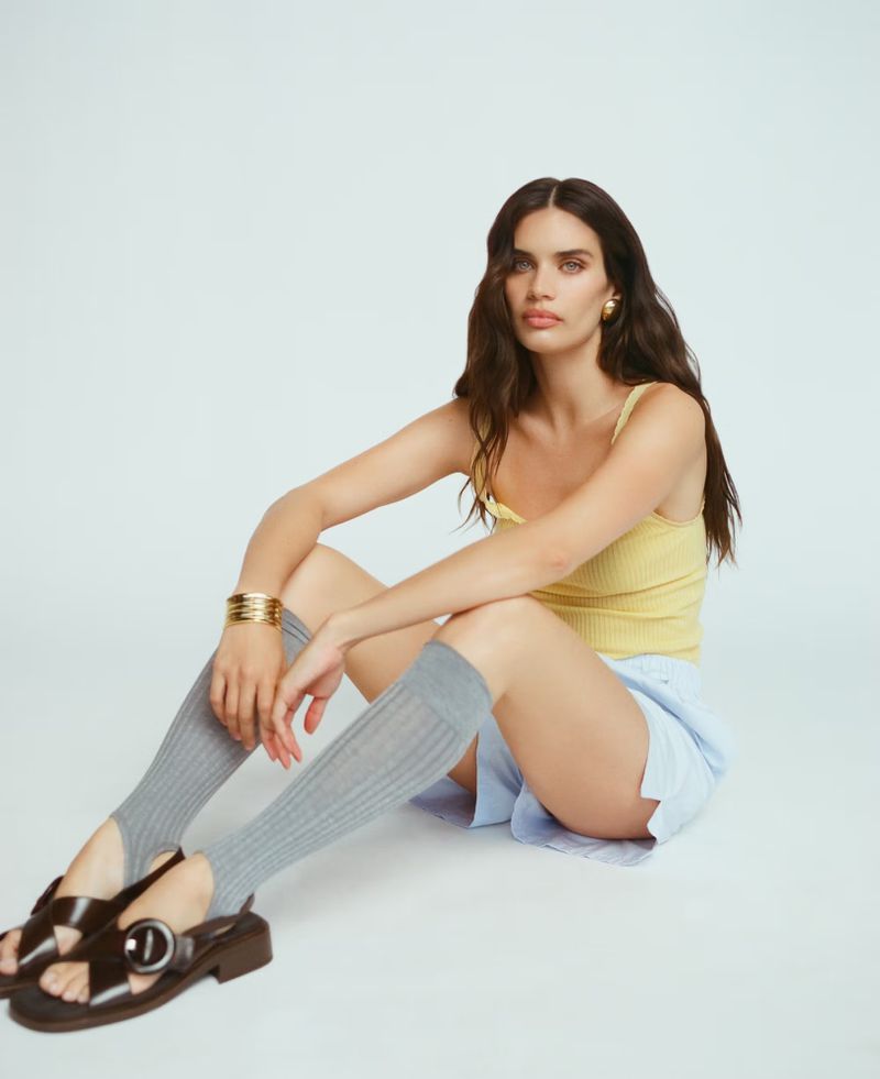 Woman in yellow tank top, blue shorts, grey leg warmers, and brown sandals poses in a studio with a neutral expression while sitting.