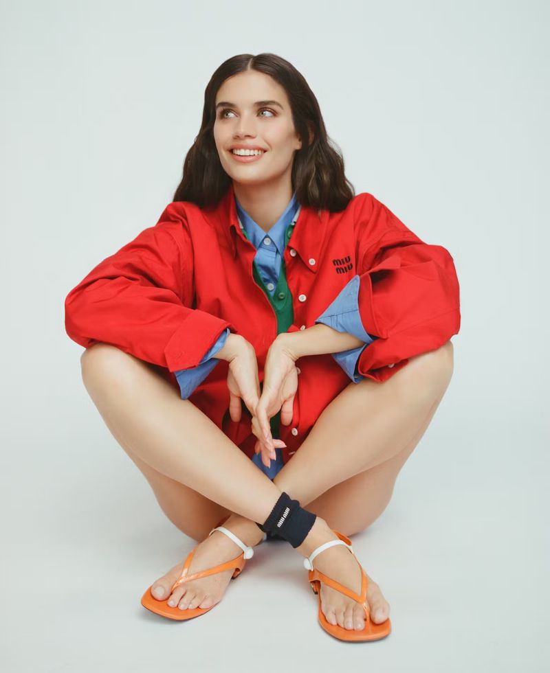 A woman in a red jacket sits cross-legged on a white floor, smiling and looking up, showcasing a relaxed yet stylish fashion aesthetic.