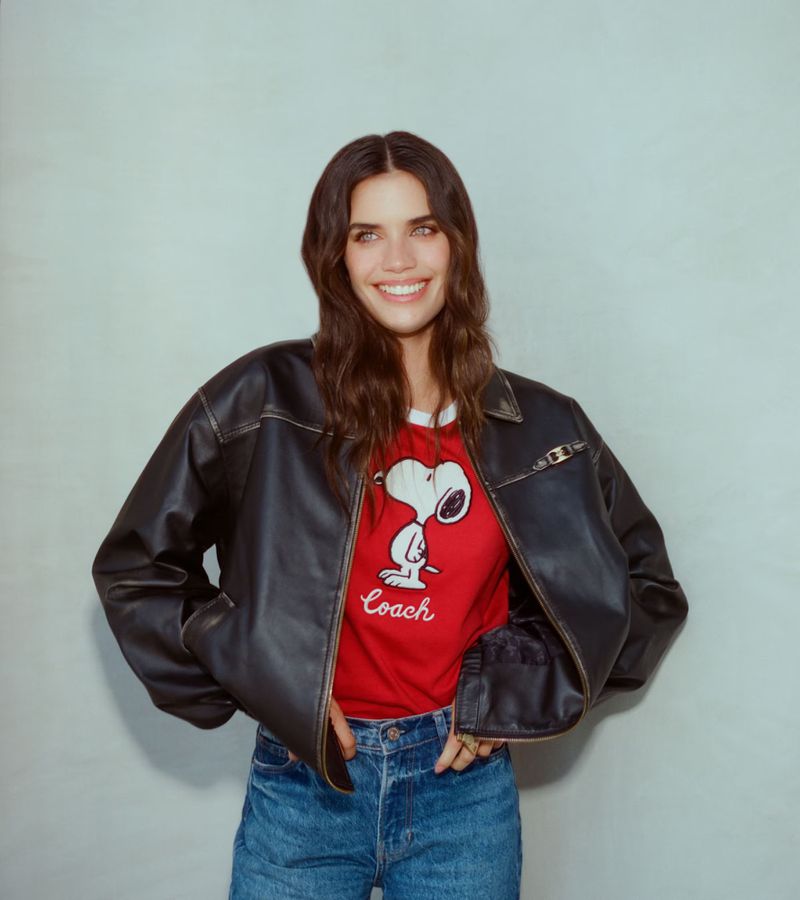 Adult woman smiling in a studio wearing a leather jacket over a red t-shirt with a cartoon, and blue denim jeans.