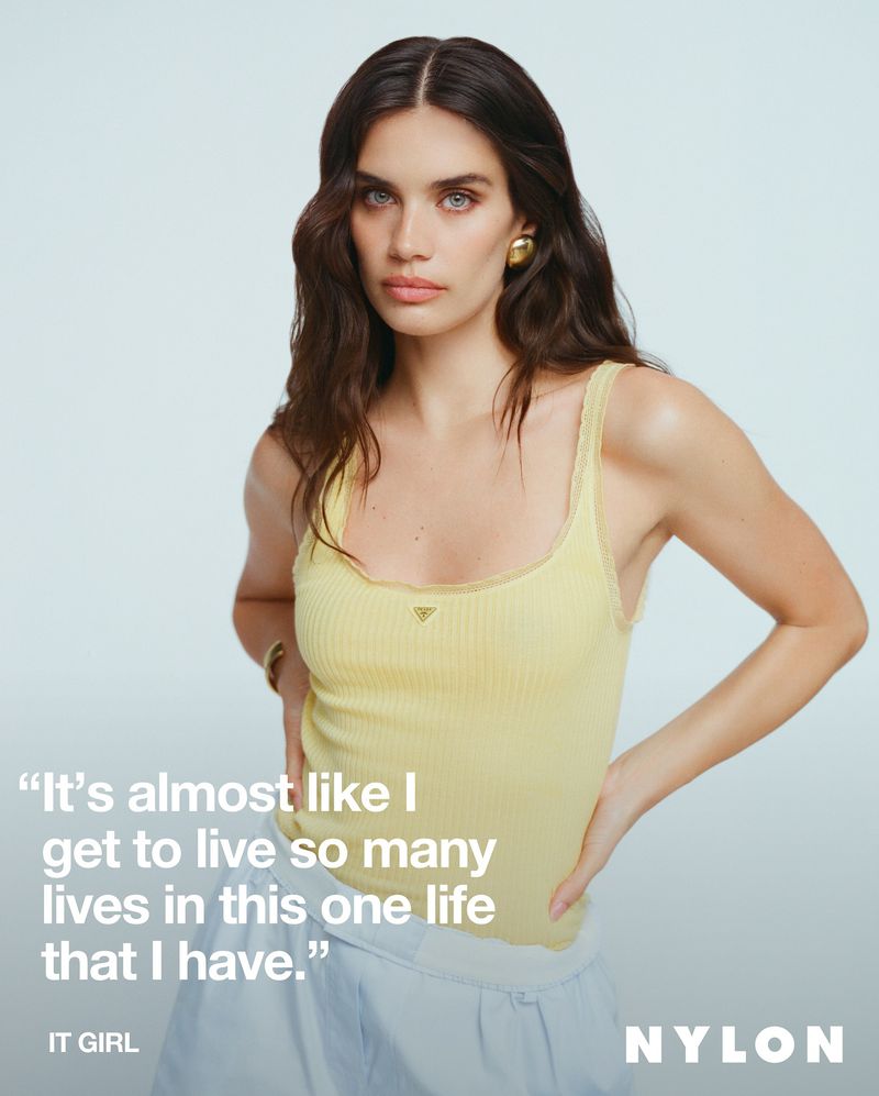 Brunette woman in yellow top poses in front of a light background, wearing gold earrings and a bracelet.  Visible text includes NYLON magazine.