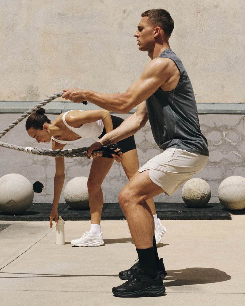 A man in athletic wear squats while training with battle ropes beside a woman in sportswear outdoors.