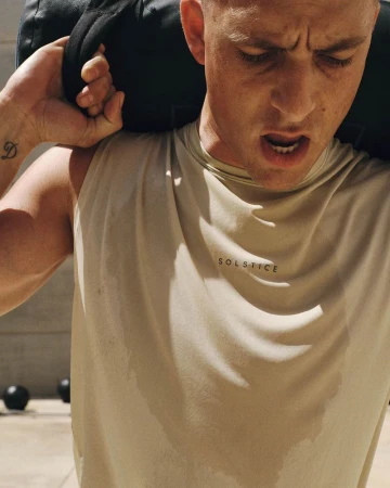 Man carrying a heavy black workout sandbag over his shoulders in a sweat-stained beige sleeveless top under bright sunlight.