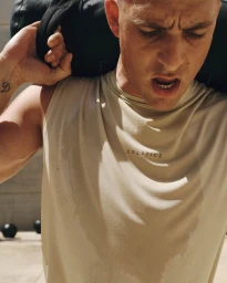 Man carrying a heavy black workout sandbag over his shoulders in a sweat-stained beige sleeveless top under bright sunlight.