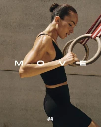 Woman in a black athletic crop top and shorts holding wooden gymnastic rings against a concrete wall.