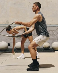 Man in a grey sleeveless shirt exercising with battle ropes alongside a woman outdoors against a concrete wall.