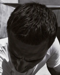 High-angle close-up of a man with short cropped dark hair looking down in a white athletic t-shirt.