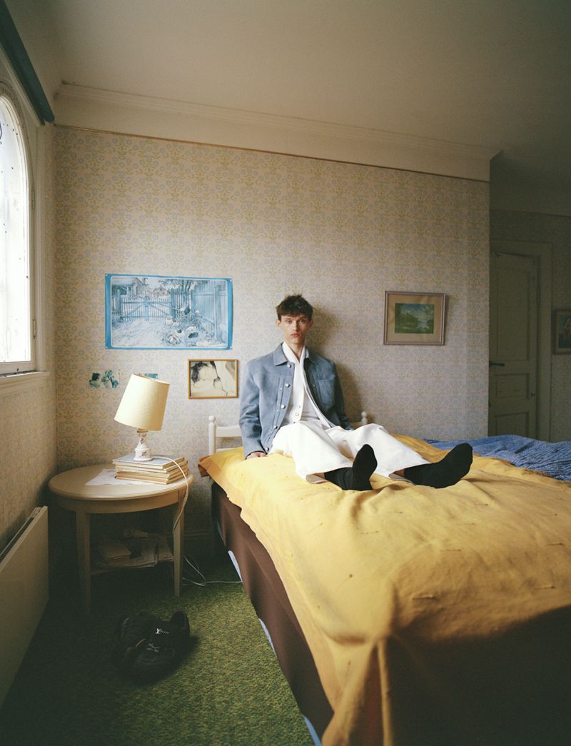 A young adult poses on a bed in an indoor setting with art on the walls, next to a round table and black shoes on the floor.