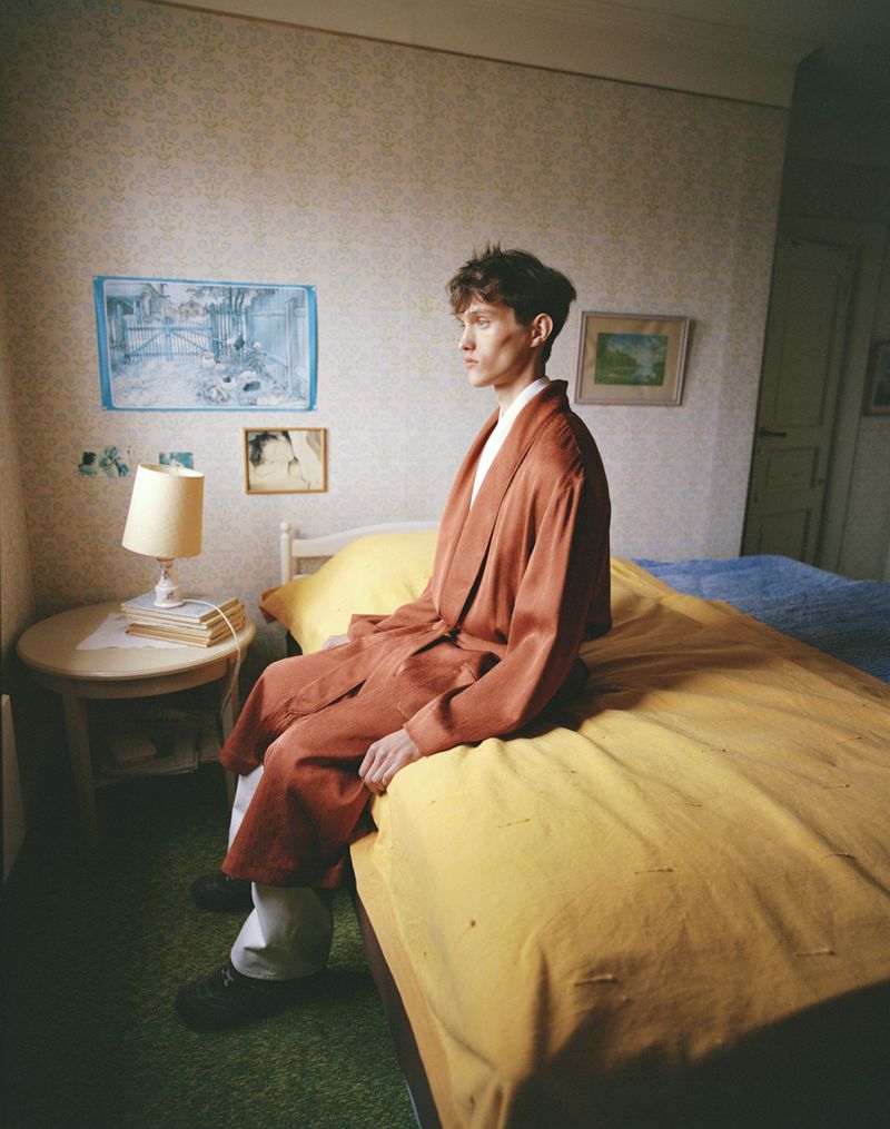 Indoor portrait of a young adult male sitting on a yellow bed wearing a rust-colored robe, looking out the window in a bedroom setting.