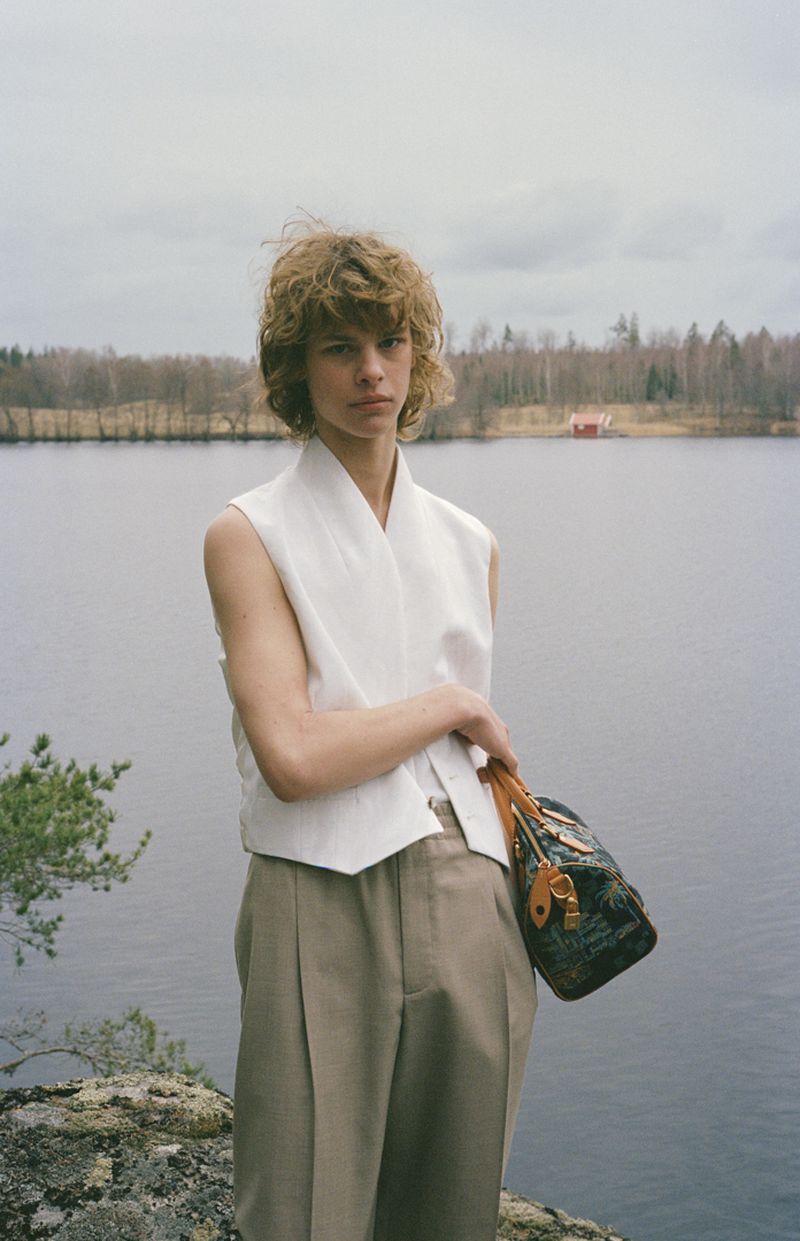 Young adult stands near a lake with a bag, wearing a vest and khaki pants, with trees and cloudy sky in the background.  