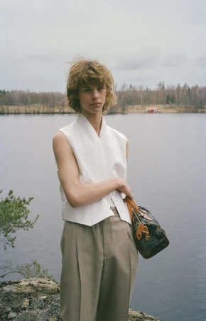 Young adult stands near a lake with a bag, wearing a vest and khaki pants, with trees and cloudy sky in the background.  
