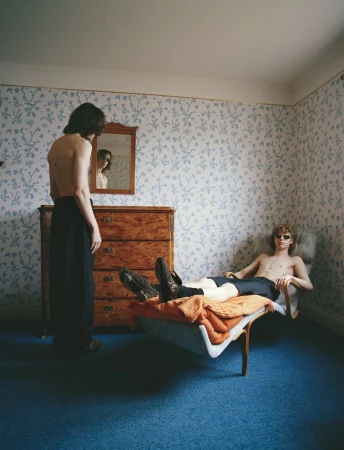 Two shirtless men pose in a bedroom with patterned wallpaper; one stands and one reclines on a chair with a blanket.
