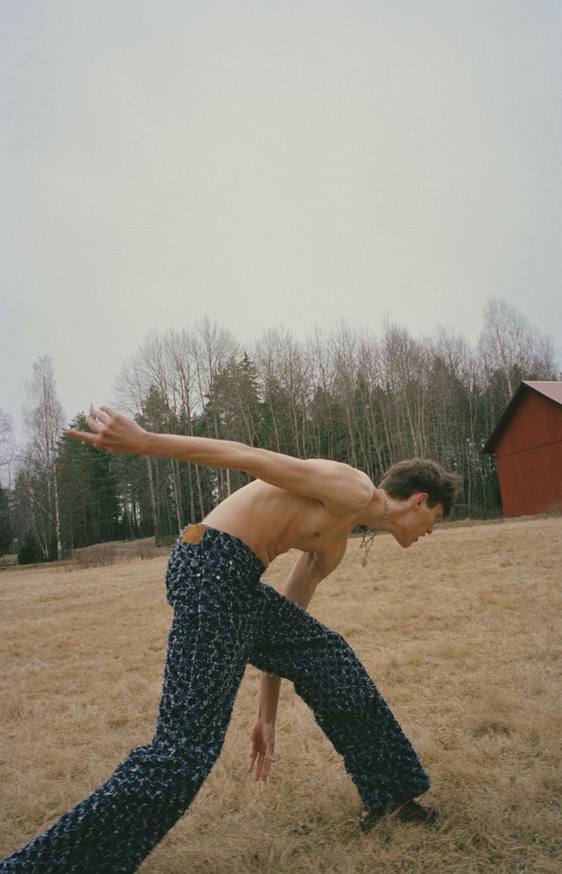 Full-body shot of a shirtless man in patterned pants crouching in a grassy field with a red barn and bare trees in the background.